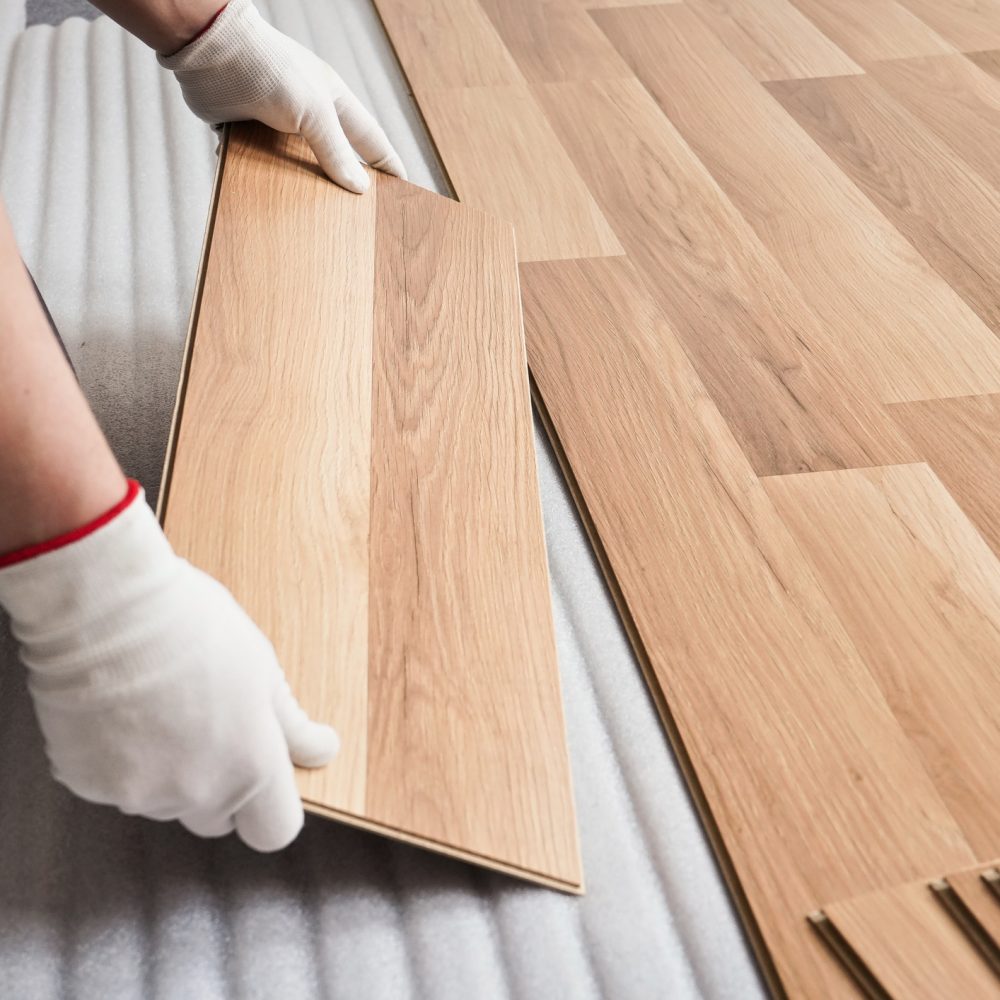 Installing laminated floor, detail on man hands with white gloves fitting wooden tile, over white foam base layer Installing laminated floor, detail on man hands with white gloves fitting wooden tile, over white foam base layer.